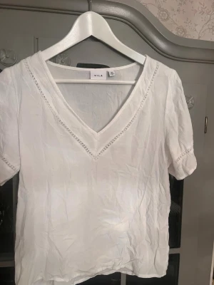 Vit v-ringad blus från Vila - Säljer en luftig vit blus från Vila i storlek 34. Blusen har korta ärmar, v-ringning med spetsdetaljer och är tillverkad i ett tunt, mjukt bomullstyg. Perfekt till jeans eller kjol för en fräsch look.