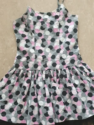 Klänning - Söt klänning med smala axelband och volangkjol. Mönstret är prickigt i svart, vit, grå och ljusrosa. Klänningen har en smockad rygg för extra komfort och en lekfull look. Perfekt för dig som gillar färg och mönster! Ena axelbandet har gått av.