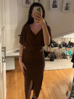 Brun midiklänning med slits - Snygg brun midiklänning med v-ringning och draperad överdel. Kjolen har rynkade detaljer framtill och en slits som ger en trendig look. Klänningen har korta, vida ärmar och markerad midja. Perfekt för dig som gillar stilrena och moderna plagg.
