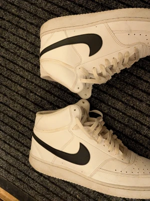 Vita Nike high top sneakers - Säljer ett par klassiska vita high top sneakers från Nike med svart swoosh på sidan. Skorna är i läder med perforerade detaljer vid tårna och har vita skosnören