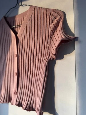 Rosa ribbad kortärmad kofta - Säljer en söt rosa ribbad kofta med korta ärmar och v-ringning. Koftan har knappar framtill och en croppad passform med vågig kant nedtill. Perfekt till sommaren och enkel att styla med jeans eller kjol.