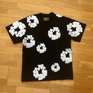 Denim Tears T shirt - Cool svart t-shirt från Denim Tears med stora vita blommor tryckta över hela plagget. T-shirten har en klassisk passform och är kortärmad, perfekt för dig som vill sticka ut med en unik och trendig stil.