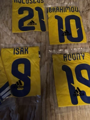 Svenska landslagströjor Adidas - Fyra gula svenska landslagströjor från Adidas med blå detaljer och spelarnamn på ryggen: Kulusevski 21, Ibrahimovic 10, Isak 9 och Roony 19. Klassisk design med stora siffror och Adidas-logga. Perfekt för dig som älskar fotboll och vill stötta Sverige.