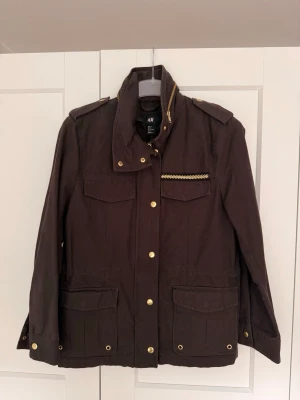 Mörkbrun trenchcoat från H&M - Stilren mörkbrun trenchcoat från H&M med guldknappar, axelklaffar och dekorativ gulddetalj vid bröstfickan. Jackan har fyra stora fickor framtill och hög krage med gulddragkedja. Tillverkad i ett slitstarkt bomullsliknande material.