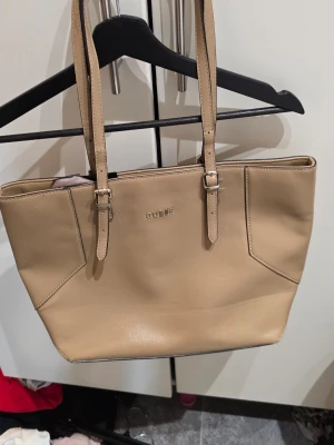 Beige handväska från Guess - Snygg beige handväska från Guess med silverfärgad logga framtill. Väskan har två långa axelband med justerbara spännen och är tillverkad i slätt konstläder. Stilren design med diskreta sömmar och rymligt huvudfack.