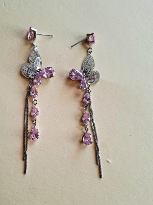 Silvriga örhängen med fjärilar och lila stenar - Unika örhängen i silverfärg med detaljerade fjärilar och flera droppformade lila stenar. Örhängena har långa kedjor som ger en cool och elegant look. Perfekta för dig som vill sticka ut och addera lite extra bling till din stil.