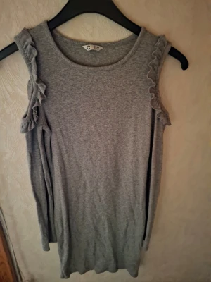 Grå ärmlös klänning med volang - Grå klänning från Cubus med ärmlös design och volangdetaljer längs axlarna. Ribbad struktur i mjukt material som ger en skön känsla och snygg passform. Enkel och stilren modell som är lätt att matcha.