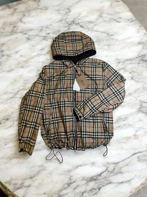 Burberry vindjacka - Snygg beige vindjacka från Burberry med klassiskt rutigt mönster i svart, vitt och rött. Jackan har huva, dragkedja framtill, svarta detaljer och justerbar dragsko i nederkant. Insidan är svart och materialet känns lätt och smidigt, perfekt för blåsiga dagar. ⚠️ Stor i storlek passar mer M ⚠️ 