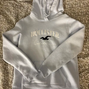 Ljusblå hoodie från Hollister - Snygg ljusblå hoodie från Hollister med broderad logga och text på bröstet. Klassisk känguruficka framtill och ribbade muddar vid ärmar och nederkant. Mjuk och skön bomullsblandning, perfekt för chill dagar. Huva med dragsko för extra stilpoäng.