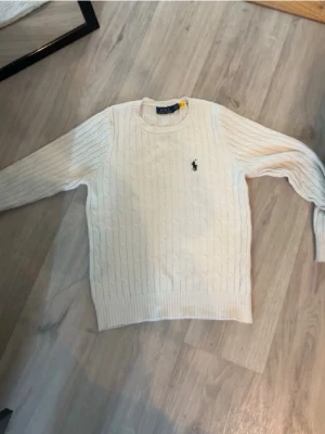 Vit kabelstickad tröja Polo Ralph Lauren - Kabelstickad vit tröja från Polo Ralph Lauren med rund halsringning och lång ärm. Klassisk design med broderad marinblå logga på bröstet. Ribbstickade muddar vid ärmslut och nederkant. Perfekt för en clean och stilren look.