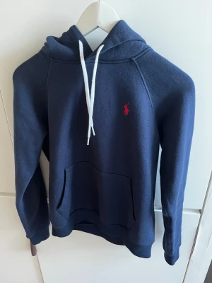 Ralph Lauren hoodie - Mörkblå hoodie från Ralph Lauren med vit dragsko i huvan och klassisk röd logga broderad på bröstet. Hoodien har känguruficka framtill och ribbade muddar vid ärmar och nederkant. Perfekt för en avslappnad och stilren look.