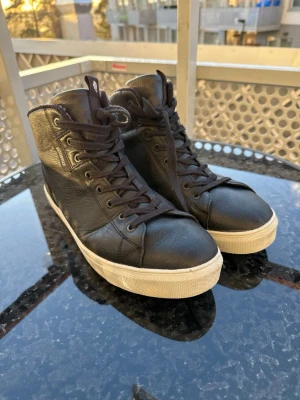Svarta high top sneakers i läder - Svarta vinter sneakers för herr, storlek 44. Använda men i gott skick.  Skorna är varma och bekväma, perfekta för kalla dagar. Stabil sula med bra grepp, vilket gör dem praktiska för vinterväglag. Klassisk svart design som är lätt att matcha med olika outfits.  Perfekta för både vardag och promenader under vintern/höst