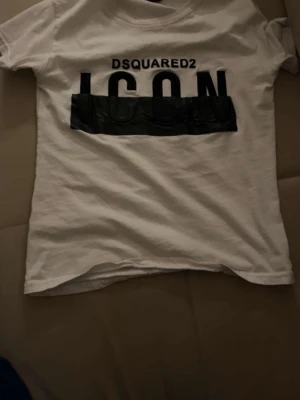 Vit Dsquared2 ICON t-shirt - Säljer en vit t-shirt från Dsquared2 med stor svart 'ICON'-text och Dsquared2-logga på bröstet. Klassisk passform och korta ärmar, perfekt till jeans eller shorts. Materialet känns mjukt och skönt mot huden.