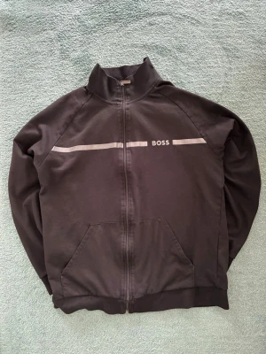 Svart ziptröja från BOSS - Svart ziptröja från BOSS med hög krage och två fickor framtill. Tröjan har en diskret silverfärgad rand och BOSS-logga över bröstet. Tillverkad i mjukt material, perfekt för en avslappnad och stilren look.