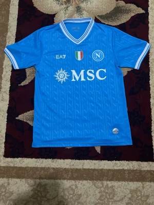 Napoli matchtröja blå EA7 - Snygg blå Napoli matchtröja från EA7 med vita detaljer och klubbmärke på bröstet. Tröjan har MSC-logga, italienska mästarmärket och rundad hals med vit-blå kant. Tillverkad i lätt och ventilerande material, perfekt för fotboll eller träning.