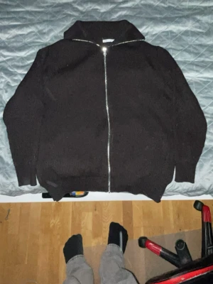 Svart stickad ziptröja med hög krage - Säljer en svart stickad tröja med dragkedja framtill och hög krage. Tröjan har långa ärmar och ribbade muddar vid ärmslut och nederkant. Perfekt för lager på lager och enkel att matcha med olika outfits.