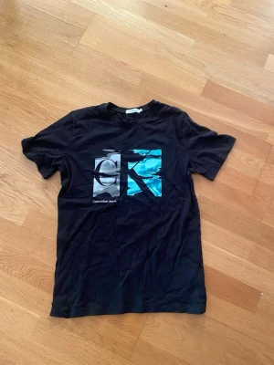 Svart Calvin Klein t-shirt med tryck - Snygg svart t-shirt från Calvin Klein Jeans med stort grafiskt CK-tryck i vitt och blått på bröstet. Klassisk passform och rund halsringning. Perfekt till jeans eller shorts för en avslappnad look.