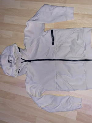 Beige hoodie från Sail Racing - Säljer en beige hoodie från Sail Racing med dragkedja framtill och en svart logga . Hoodien har en huva med dragsko och en praktisk bröstficka med dragkedja. Perfekt för dig som gillar stilren och sportig design.