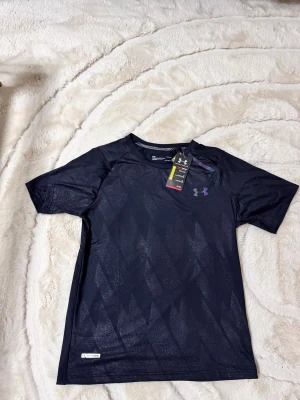 Svart träningströja Under Armour - Snygg svart träningströja från Under Armour med diskret diamantmönster och regnbågsskiftande logga på bröstet. Tillverkad i ett lätt och snabbtorkande funktionsmaterial som passar perfekt till träning och sport.