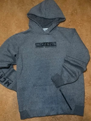 Grå hoodie från McKenzie - Snygg grå hoodie från McKenzie med tryckt logga på bröstet och textdetalj på ena ärmen. Hoodien har en klassisk huva och en praktisk ficka med dragkedja framtill. Tillverkad i mjukt sweatshirtmaterial som är perfekt för en avslappnad stil.