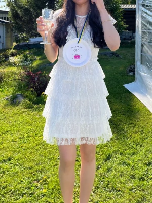 Vit spetsklänning med volanger - Säljer en superfin vit kortklänning i spets med flera lager volanger. Klänningen har ärmlös design med spetsdetaljer och ger en romantisk känsla. Perfekt till studenten!