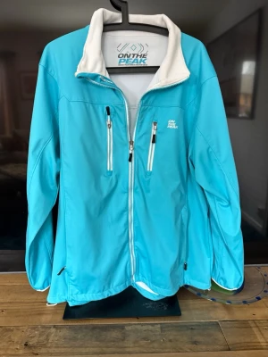Turkos vindjacka från On The Peak - Snygg turkos vindjacka från On The Peak med vit dragkedja och vita detaljer. Jackan har två bröstfickor med dragkedja, hög krage med vit insida och hel dragkedja framtill. Perfekt för blåsiga dagar och outdoor-aktiviteter.