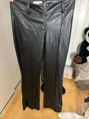 Svarta fuskskinnbyxor med dragkedja - Säljer ett par svarta byxor i fuskskinn med raka ben och medelhög midja. Byxorna har en synlig dragkedja framtill och breda hällor för bälte. Perfekta för en edgy och trendig look. Knappt använda 