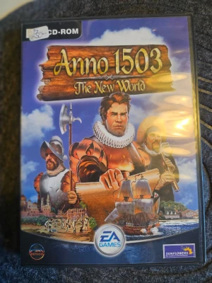 Anno 1503 The New World PC-spel - Strategispel för PC där du utforskar och bygger upp nya civilisationer i en historisk miljö. Omslaget har färgstarka illustrationer med skepp, soldater och upptäcktsresande. Spelet levereras i plastfodral och är från EA Games och Sunflowers.