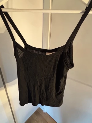 Svart ribbad linnetopp - Säljer en svart ribbad linnetopp med smala axelband. Toppen har en enkel och stilren design som passar perfekt till jeans eller kjol. Materialet är mjukt och stretchigt för en bekväm passform.