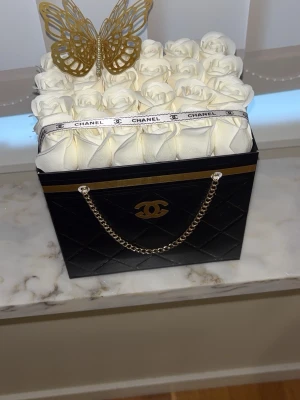 Evighetsrosor I Present väska.  - Snygg svart dekorväska med guldfärgad kedja med  C logga framtill. Väskan är fylld med vita konstrosor och dekorerad med ett vitt band samt en guldfärgad fjäril på toppen. Perfekt som unik inredningsdetalj eller present.