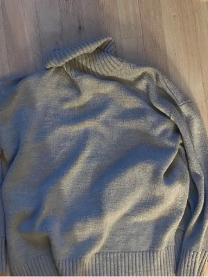 Grå polotröja i mjuk stickad kvalitet - Säljer en enkel och stilren grå stickad polotröja med bred ribbad mudd vid ärmslut och nederkant. Tröjan har en hög polokrage och är gjord i ett mjukt material som känns skönt mot huden. Perfekt för lager på lager under kyliga dagar.