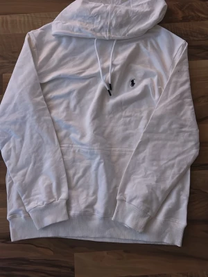 Vit hoodie från Polo Ralph Lauren - Säljer en stilren vit hoodie från Polo Ralph Lauren med klassisk huva och dragsnören. Tröjan har en broderad logga i svart på bröstet och en stor magficka. Tillverkad i mjukt bomullsmaterial och har ribbade muddar vid ärmslut och nederkant.