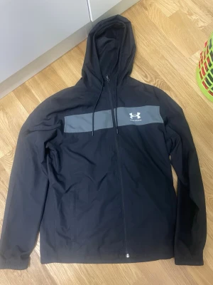 Svart vindjacka Under Armour - Svart vindjacka från Under Armour med grå panel över bröstet och vit logga. Jackan har huva med dragsko, hel dragkedja framtill och två sidofickor. Tillverkad i lätt, vindtätt material med meshfoder för extra ventilation.