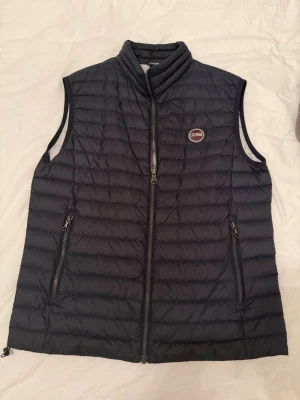 Mörkblå dunväst från Colmar - Snygg mörkblå dunväst från Colmar med quiltad design och hög krage. Västen har två dragkedjeförsedda fickor framtill och en diskret logga på bröstet. Insidan är ljusgrå och västen stängs med dragkedja. Perfekt för lager-på-lager och streetstyle. Köpt ny för ca 1900kr använd fåtal gånger 