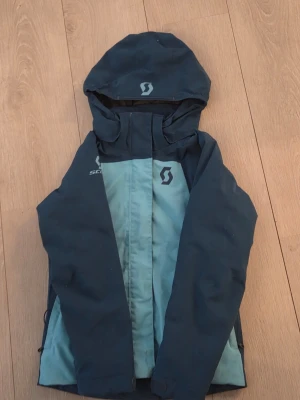 Blå vindjacka från Scott med huva - Snygg tvåfärgad vindjacka från Scott i mörkblått och ljusblått. Jackan har en praktisk huva, dragkedja framtill och logga på bröstet och huvan. Perfekt för blåsiga dagar och outdoor-aktiviteter. Tillverkad i slitstarkt syntetmaterial.