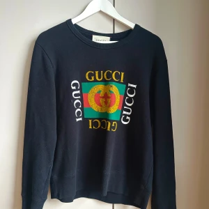 Svart sweatshirt från Gucci - Svart sweatshirt från Gucci med färgglad logga i gult, grönt och rött framtill. Tröjan har rund halsringning och långa ärmar. Tillverkad i mjukt material och har en klassisk passform. Snygg detalj med flera Gucci-tryck på bröstet.