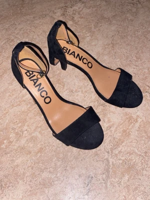 Svarta pumps med ankelrem från Bianco - Stilrena svarta pumps från Bianco med öppen tå och bred rem över foten. Skorna har en smal ankelrem med spänne och en medelhög klack. Utsidan är i mockaimitation och innersulan är beige.