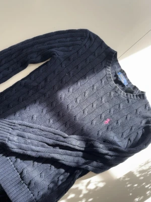 Mörkblå kabelstickad tröja Polo Ralph Lauren - Kabelstickad mörkblå tröja från Polo Ralph Lauren med rund halsringning och rosa broderad logga på bröstet. Tröjan har långa ärmar och ribbade muddar vid ärmslut och nederkant. Perfekt för lager på lager och klassisk stil.