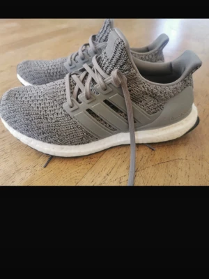 Grå Adidas Ultra Boost sneakers - Snygga Adidas Ultra Boost sneakers i grått med stickad ovandel och klassiska tre ränder på sidan. Skorna har en vit Boost-sula för extra komfort och gråa snören. Perfekta för dig som gillar stilrena och bekväma sneakers.