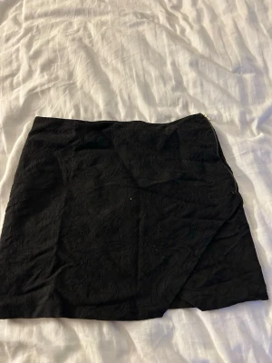 Svart omlottkjol med diskret mönster - Kort svart omlottkjol med subtilt mönster och dragkedja i sidan. Kjolen har en stilren look och passar perfekt till många olika outfits. Materialet känns mjukt och följsamt.