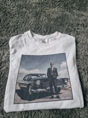 T-shirt med tryck - Säljer en vit t-shirt från L'Arte med ett stort tryck framtill som visar en person i kostym framför en klassisk bil. T-shirten har rund hals och korta ärmar. Materialet är mjuk bomull och passformen är normal. Men ser en liten defekt vid kragen men den märks inte när man bär den och de syns knappt.