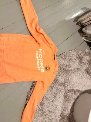 Orange stickad tröja Morris Company - Snygg orange stickad tröja från Morris Company med vit logotyptext på bröstet och ett broderat emblem. Tröjan har rund halsringning, långa ärmar och ribbade muddar vid ärmslut och nederkant. #sthlm #stickad #cool #stockholmsstil