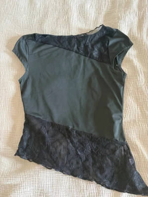 Svart topp med spetsdetaljer - Snygg svart topp med asymmetrisk design och spetsdetaljer både upptill och nedtill. Kortärmad modell med mjukt, stretchigt material och transparent spets som ger en cool kontrast. Perfekt för dig som gillar unika detaljer och stilren look.
