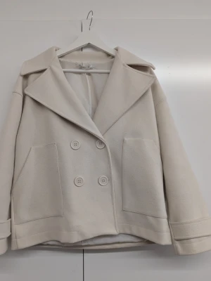 Beige kappa från design by Si - Trendig kappa från design by si. Stilren kort kappa i beige med dubbelknäppning och stora utanpåfickor. Klassisk krage och breda slag ger en trendig look. Perfekt för lager-på-lager och enkel att matcha. Troligtvis i ullblandning för extra värme. Storlek XS/S