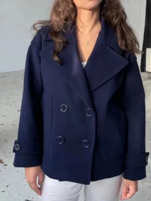 Design by si kappa - Kort mörkblå rock med dubbelknäppning och bred krage. Rocken har fyra stora knappar framtill, två sidofickor och dekorativa knappar vid ärmsluten. Tillverkad i ullblandning för en klassisk och stilren look. En av banden vid armén har lossnat så den hänger löst men går å sy på jätte enkelt