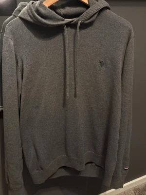 Mörkgrå hoodie från U.S. Polo Assn. - Snygg mörkgrå hoodie från U.S. Polo Assn. med broderad logga på bröstet. Tröjan har huva med dragsko och ribbade muddar vid ärmslut och nederkant. Har ett hål vid luvan se bild.Storlek L, skriv vid frågor eller funderingar!
