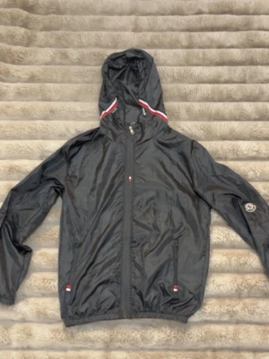 Svart vindjacka med huva  - Snygg svart vindjacka  med huva och dragkedja framtill. Jackan har vita och röda detaljer vid huvan och små loggor på ärmen och vid fickorna. Tillverkad i ett glansigt, lätt material som passar perfekt för blåsiga dagar. Köpt från Plick men säljer vidare då den var för liten för mig :) 