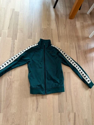Grön track jacket från Kappa XS - Snygg grön track jacket från Kappa med dragkedja och vita ränder med Kappas logga längs ärmarna. Jackan har hög krage och muddar vid ärmslut och midja. Perfekt för sport eller streetwear-stil.