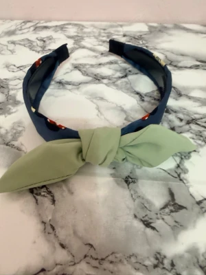 Blå hårband med grön rosett - Snyggt hårband i blått tyg med en stor ljusgrön rosett framtill. Hårbandet har en vadderad båge och är dekorerad med små röda och vita detaljer på sidorna. Perfekt accessoar för att lyfta din look med en färgklick.
