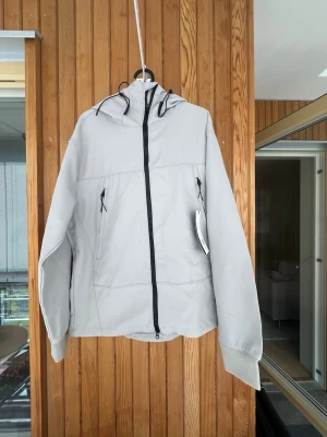 Ljusgrå vindjacka med huva - Snygg ljusgrå vindjacka med hel dragkedja, två fickor med dragkedja och justerbar huva. Jackan har en minimalistisk design och är tillverkad i ett lätt material som passar perfekt för blåsiga dagar. 